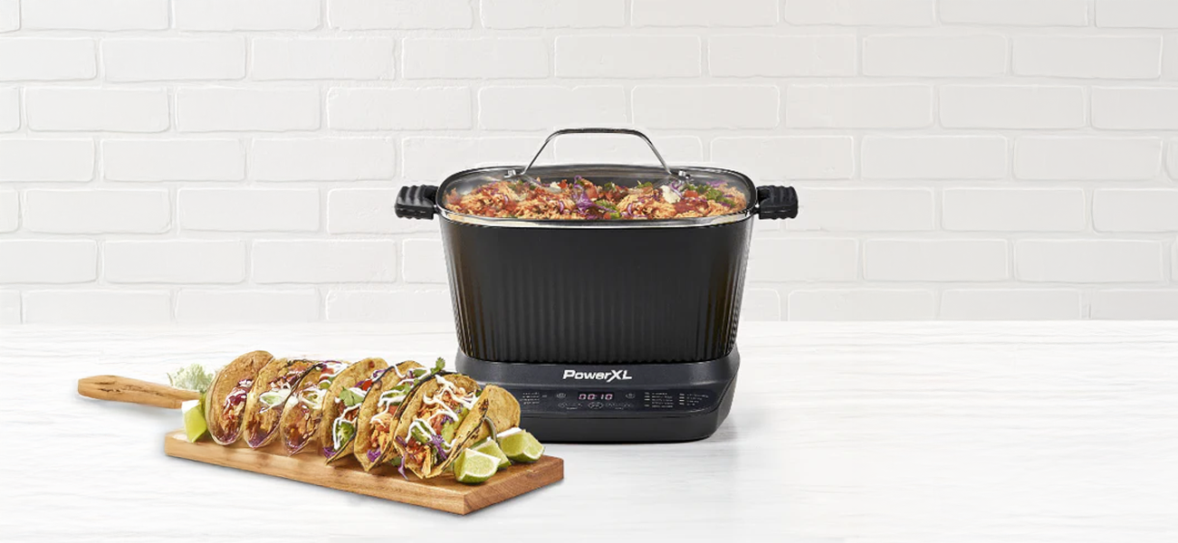 Powerxl Stimax multi cooker with food on a white surface, next to a wooden board with tacos.